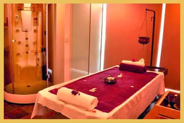 A single treatment room with a massage table dressed in deep maroon linen and folded white towels topped with flower petals, beside a glass-walled steam shower cabin and a hanging oil burner.