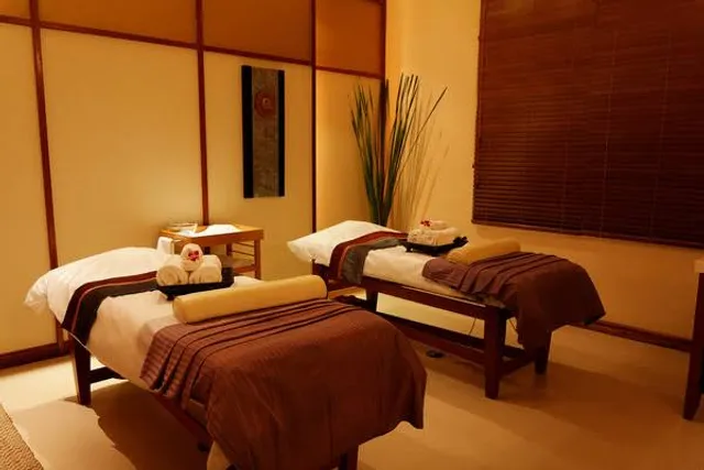 A twin treatment room with two massage tables set side by side in white and brown linens, each with folded towels and flower petals, in front of a wood-slat partition and wooden blinds.